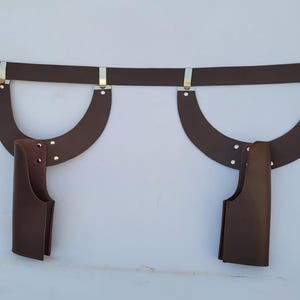 Puede incluir: Un cintur&oacute;n de cuero marr&oacute;n con dos piezas de cuero curvadas adjuntas y dos bolsas de cuero marr&oacute;n. El cintur&oacute;n tiene una hebilla plateada y detalles met&aacute;licos plateados. Las bolsas est&aacute;n unidas a las piezas de cuero curvadas con remaches.