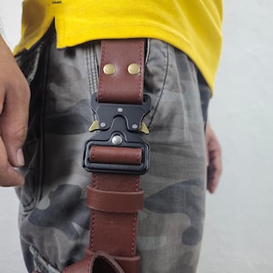 May include: A brown leather belt with a black metal buckle and gold-colored accents. The belt is attached to camouflage pants and a yellow shirt. The buckle is a prominent feature, with a secure, modern design.