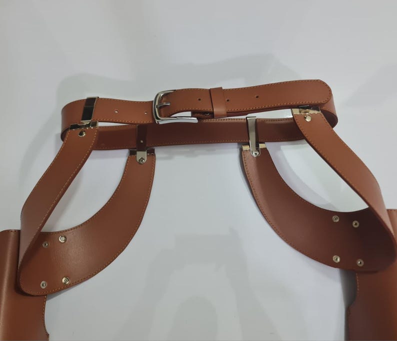 Puede incluir: Un cintur&oacute;n arn&eacute;s de cuero marr&oacute;n claro con una correa central en la cintura y una hebilla de metal plateado. El dise&ntilde;o incluye paneles laterales anchos y curvos conectados al cintur&oacute;n principal mediante herrajes y remaches plateados.
