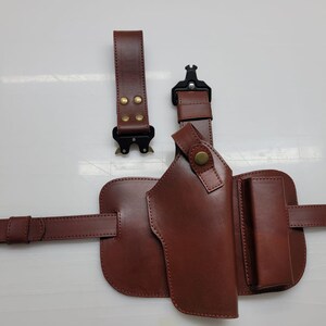 May include: A brown leather holster with a belt and two detachable straps. The holster has a pouch and a magazine holder. The straps have black metal buckles and brass rivets. The leather is a rich, dark brown color.