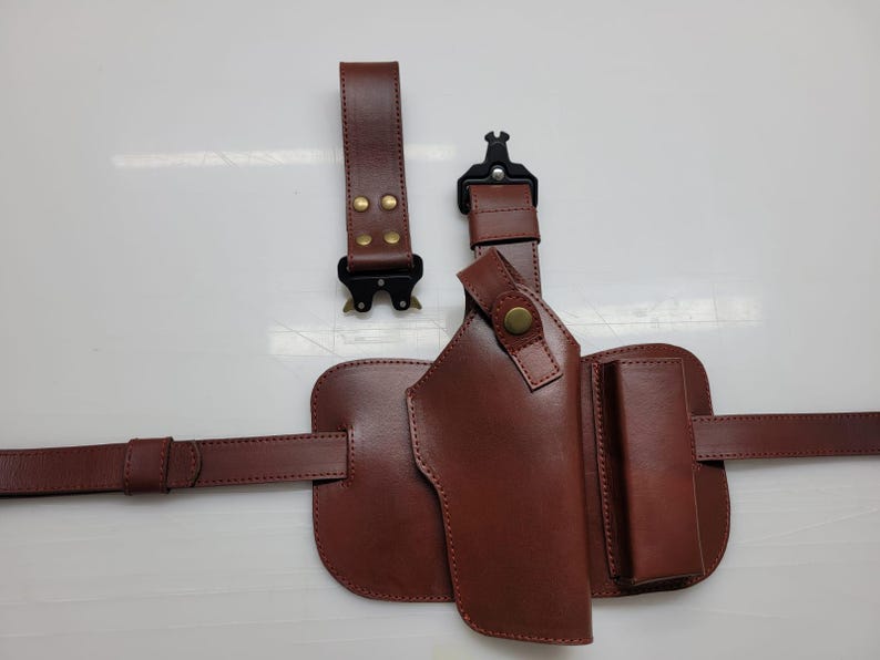 May include: A brown leather holster with a belt and two detachable straps. The holster has a pouch and a magazine holder. The straps have black metal buckles and brass rivets. The leather is a rich, dark brown color.
