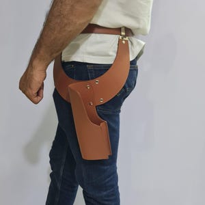 Puede incluir: Una funda de cuero color canela sujeta a un cintur&oacute;n de cuero a juego. La funda presenta un dise&ntilde;o de panel ancho y curvo que se envuelve alrededor de la cadera, asegurado con remaches met&aacute;licos. La estructura es r&iacute;gida y abierta por arriba.