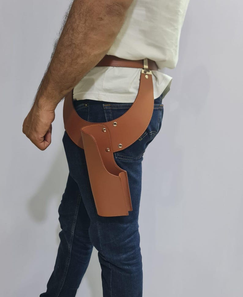 Puede incluir: Una funda de cuero color canela sujeta a un cintur&oacute;n de cuero a juego. La funda presenta un dise&ntilde;o de panel ancho y curvo que se envuelve alrededor de la cadera, asegurado con remaches met&aacute;licos. La estructura es r&iacute;gida y abierta por arriba.