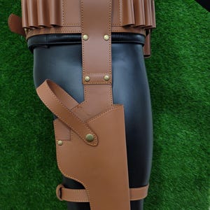 May include: A brown leather holster and belt set. The holster is attached to a leg strap and features a curved design. The belt has multiple loops and is secured with a buckle. The set is made of brown leather and has brass-colored rivets.