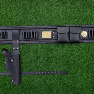 May include: Black leather utility belt with a holster, pouches, and cartridge holders. The belt features gold-colored accents, including medallions and a rectangular buckle. The belt is laid out on a green surface.