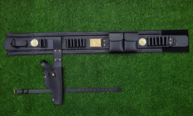 May include: Black leather utility belt with a holster, pouches, and cartridge holders. The belt features gold-colored accents, including medallions and a rectangular buckle. The belt is laid out on a green surface.