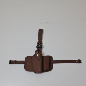 May include: A brown leather shoulder holster with a vertical strap, a holster for a firearm, and a pouch. The holster has a buckle and adjustable straps. The background is a plain white wall.