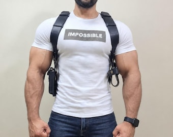 May include: A white short-sleeved t-shirt with the word IMPOSSIBLE printed in a rectangular box, worn with a black leather shoulder holster harness, a black baseball cap, and dark blue denim jeans, accessorized with a black digital watch.