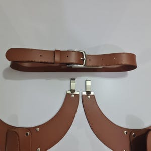 Puede incluir: Un cintur&oacute;n de cuero marr&oacute;n con una hebilla de metal plateado colocado sobre dos accesorios curvos de cuero marr&oacute;n en forma de funda. Cada accesorio cuenta con clips de metal plateado en la parte superior y remaches plateados en los bordes.