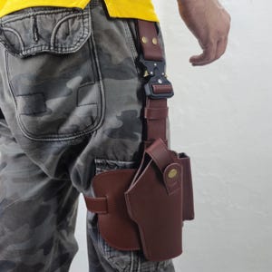 May include: A brown leather holster with a black buckle and strap, designed to be worn on the leg. The holster is attached to a belt loop and features a secure design for carrying a firearm. The image shows the holster attached to a person's leg.