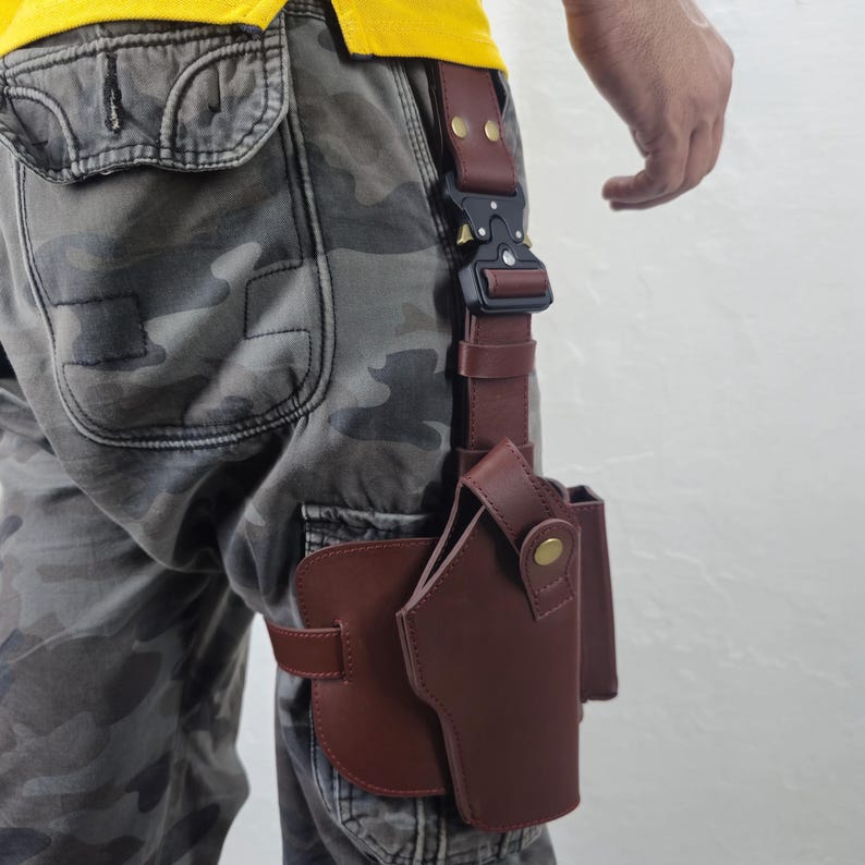 May include: A brown leather holster with a black buckle and strap, designed to be worn on the leg. The holster is attached to a belt loop and features a secure design for carrying a firearm. The image shows the holster attached to a person's leg.