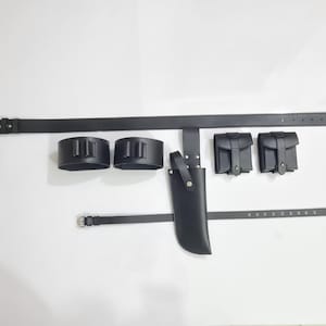 May include: A black leather belt with a rectangular buckle, two cartridge holders, two small pouches, a knife sheath, and a smaller belt. The items are arranged on a white surface.