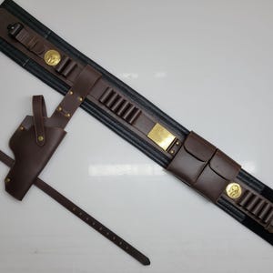 May include: A brown leather gun belt with a holster and a separate belt. The belt has cartridge loops, pouches, and gold-colored metal accents. The holster is designed to hold a handgun.