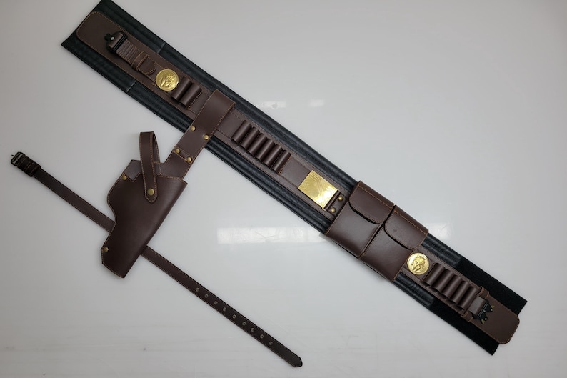 May include: A brown leather gun belt with a holster and a separate belt. The belt has cartridge loops, pouches, and gold-colored metal accents. The holster is designed to hold a handgun.