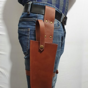 May include: A brown leather thigh holster with a belt loop and a strap that goes around the leg. The holster has a flap that covers the top of the holster and a snap closure. The holster is worn on a person wearing blue jeans and a plaid shirt.