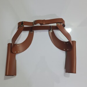 Puede incluir: Un cintur&oacute;n de doble funda de cuero marr&oacute;n que presenta una correa de cintura con una hebilla de metal plateado y dos fundas laterales verticales unidas a paneles de cuero curvos. El conjunto est&aacute; asegurado con remaches y herrajes de metal plateado.