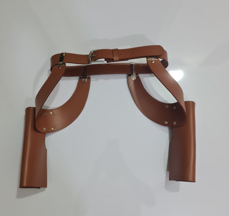 Puede incluir: Un cintur&oacute;n de doble funda de cuero marr&oacute;n que presenta una correa de cintura con una hebilla de metal plateado y dos fundas laterales verticales unidas a paneles de cuero curvos. El conjunto est&aacute; asegurado con remaches y herrajes de metal plateado.