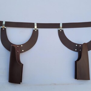 Puede incluir: Un cintur&oacute;n de cuero marr&oacute;n con hebilla plateada y detalles met&aacute;licos. Dos piezas de cuero curvadas est&aacute;n unidas al cintur&oacute;n, cada una con una funda de cuero marr&oacute;n. El cintur&oacute;n est&aacute; dise&ntilde;ado para usarse alrededor de la cintura.