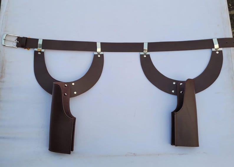 Puede incluir: Un cintur&oacute;n de cuero marr&oacute;n con hebilla plateada y detalles met&aacute;licos. Dos piezas de cuero curvadas est&aacute;n unidas al cintur&oacute;n, cada una con una funda de cuero marr&oacute;n. El cintur&oacute;n est&aacute; dise&ntilde;ado para usarse alrededor de la cintura.