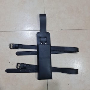 May include: Black leather sheath with two adjustable straps and buckles. The sheath has a vertical design with a loop at the top and is designed to be worn on the leg. The straps are secured with silver-colored buckles.