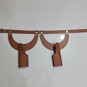 Puede incluir: Un cintur&oacute;n de cuero color canela con dos accesorios de funda sim&eacute;tricos en forma de media luna. El cintur&oacute;n incluye una hebilla de metal plateado y herrajes a juego, con fundas verticales aseguradas con remaches a los paneles curvos.