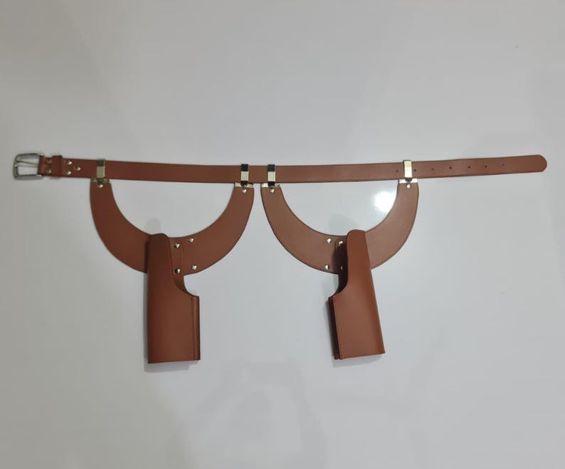 Puede incluir: Un cintur&oacute;n de cuero color canela con dos accesorios de funda sim&eacute;tricos en forma de media luna. El cintur&oacute;n incluye una hebilla de metal plateado y herrajes a juego, con fundas verticales aseguradas con remaches a los paneles curvos.