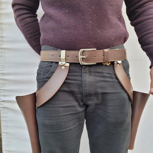 Puede incluir: Cintur&oacute;n de cuero marr&oacute;n para funda de pistola con dos bolsas adjuntas. El cintur&oacute;n tiene una hebilla plateada y dos clips plateados. La persona lleva un jersey morado oscuro y vaqueros negros.