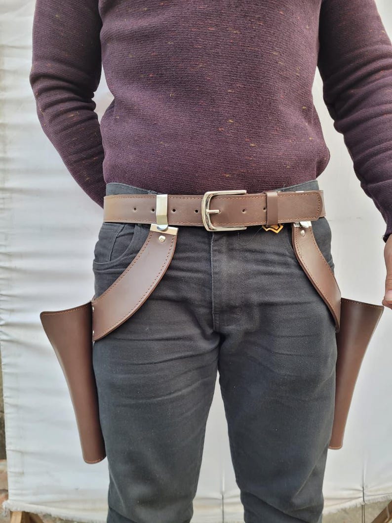 Puede incluir: Cintur&oacute;n de cuero marr&oacute;n para funda de pistola con dos bolsas adjuntas. El cintur&oacute;n tiene una hebilla plateada y dos clips plateados. La persona lleva un jersey morado oscuro y vaqueros negros.