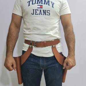 Puede incluir: Una camiseta blanca de manga corta con el texto azul TOMMY JEANS y un peque&ntilde;o logotipo de bandera, combinada con vaqueros azul oscuro y un cintur&oacute;n de cuero marr&oacute;n con dos fundas laterales estilo pistolera con herrajes de metal plateado.