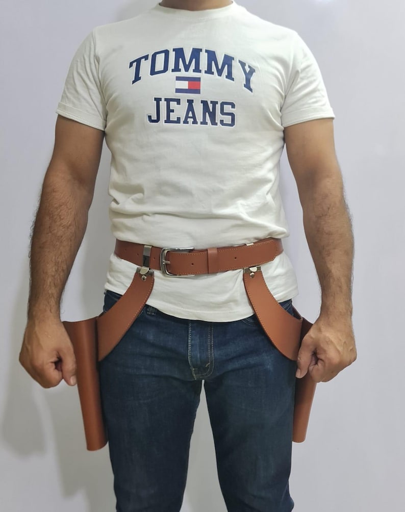 Puede incluir: Una camiseta blanca de manga corta con el texto azul TOMMY JEANS y un peque&ntilde;o logotipo de bandera, combinada con vaqueros azul oscuro y un cintur&oacute;n de cuero marr&oacute;n con dos fundas laterales estilo pistolera con herrajes de metal plateado.