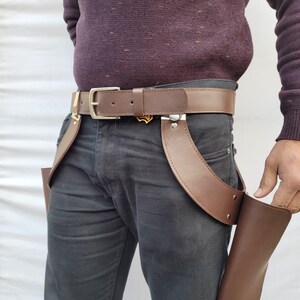 Puede incluir: Cintur&oacute;n de cuero marr&oacute;n con hebilla plateada y dos pistoleras de cuero marr&oacute;n adjuntas. El cintur&oacute;n se usa con pantalones grises oscuros y un su&eacute;ter burdeos. Las pistoleras est&aacute;n dise&ntilde;adas para guardar objetos.