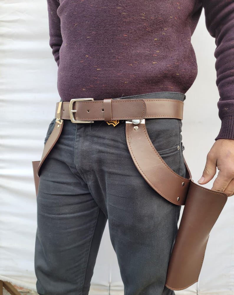 Puede incluir: Cintur&oacute;n de cuero marr&oacute;n con hebilla plateada y dos pistoleras de cuero marr&oacute;n adjuntas. El cintur&oacute;n se usa con pantalones grises oscuros y un su&eacute;ter burdeos. Las pistoleras est&aacute;n dise&ntilde;adas para guardar objetos.