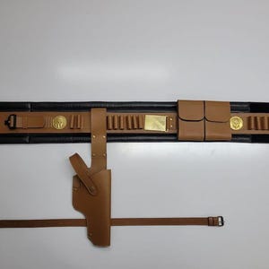 May include: A brown leather gun belt with a holster, cartridge holders, and a separate belt. The belt has a black backing and gold-colored accents, including a buckle and decorative medallions. The holster is designed to hold a handgun.