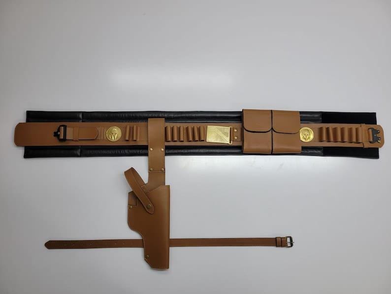 May include: A brown leather gun belt with a holster, cartridge holders, and a separate belt. The belt has a black backing and gold-colored accents, including a buckle and decorative medallions. The holster is designed to hold a handgun.