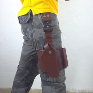 May include: A brown leather holster with a magazine pouch attached, secured to a belt with a black buckle. The holster is designed to be worn on the leg. The person is wearing camouflage pants and a yellow shirt.