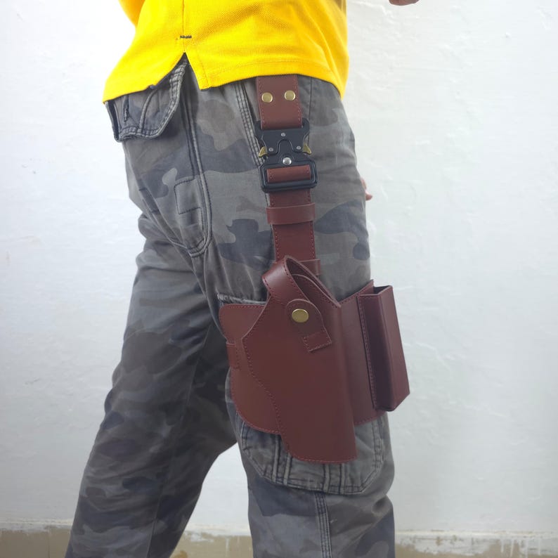 May include: A brown leather holster with a magazine pouch attached, secured to a belt with a black buckle. The holster is designed to be worn on the leg. The person is wearing camouflage pants and a yellow shirt.
