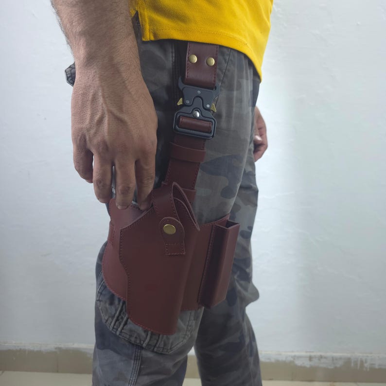 May include: A brown leather thigh holster with a matching belt and a black metal buckle. The holster has a pouch and a magazine holder. The belt is secured with two rivets.