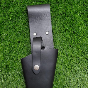 May include: A black leather sheath with a belt loop and a silver-colored snap closure. The sheath is designed to hold a small tool or knife. The background is green.