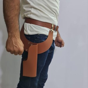 Puede incluir: Una funda de cuero color canela sujeta a un cintur&oacute;n de cuero a juego con una hebilla de metal plateado. La funda presenta un dise&ntilde;o curvo con remaches plateados visibles que aseguran la estructura de cuero en la cadera y el muslo.