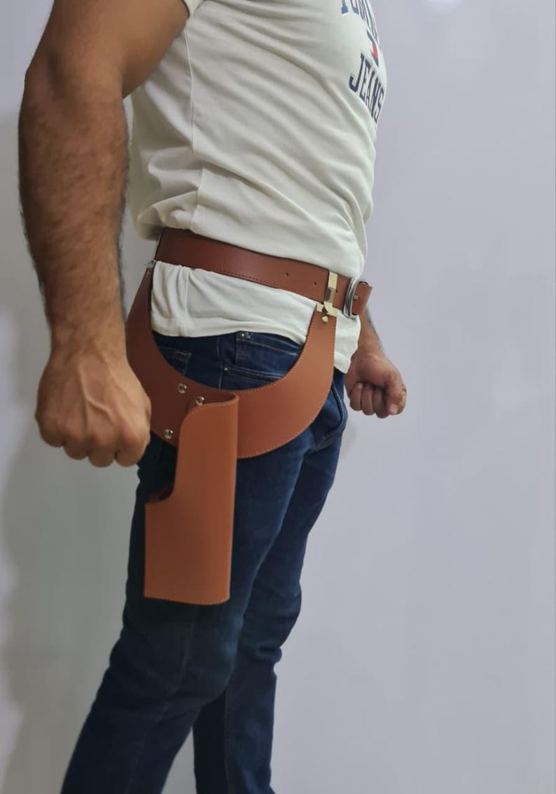 Puede incluir: Una funda de cuero color canela sujeta a un cintur&oacute;n de cuero a juego con una hebilla de metal plateado. La funda presenta un dise&ntilde;o curvo con remaches plateados visibles que aseguran la estructura de cuero en la cadera y el muslo.