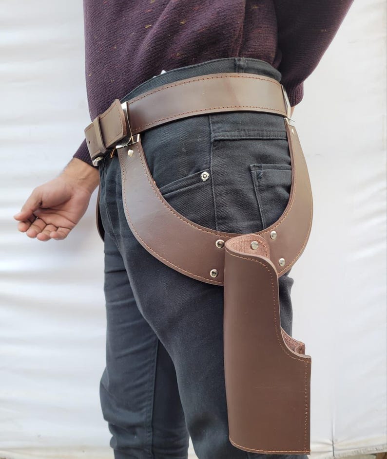 Puede incluir: Juego de cintur&oacute;n y funda de pistola de cuero marr&oacute;n. El cintur&oacute;n es de color marr&oacute;n medio con una hebilla plateada. La funda est&aacute; sujeta al cintur&oacute;n y tiene una pieza de soporte curvada. El conjunto est&aacute; dise&ntilde;ado para llevarse en la cadera.