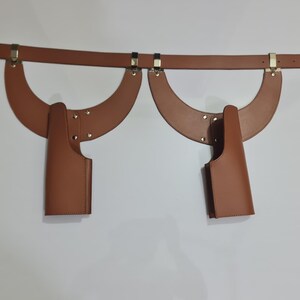 Puede incluir: Un arn&eacute;s de cuero color canela con un cintur&oacute;n horizontal conectado a dos piezas de hombro sim&eacute;tricas en forma de media luna. Cada lado incluye un accesorio de funda vertical asegurado con remaches met&aacute;licos y herrajes dorados.