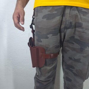 May include: A brown leather thigh holster with a belt and a phone case. The holster is attached to a belt that wraps around the thigh. The person is wearing camouflage pants and a yellow shirt.