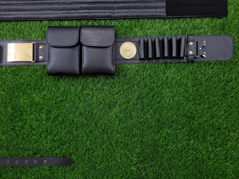 May include: Black leather utility belt with two pouches, cartridge holders, and a gold-colored buckle. The belt features a gold-colored emblem and is set against a green background.