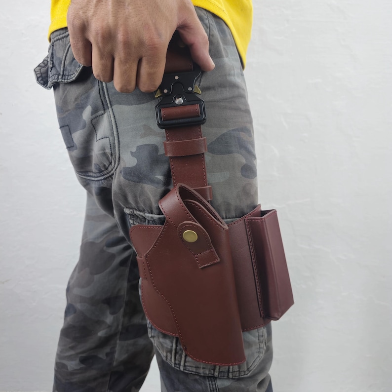 May include: A brown leather holster with a matching magazine pouch attached. The holster is secured to the leg with a brown leather strap and a black metal buckle. The person is wearing camouflage pants and a yellow shirt.
