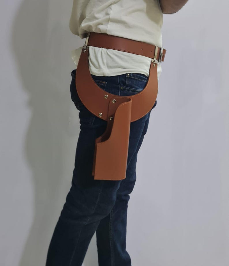 Puede incluir: Un cintur&oacute;n de cuero color canela con una funda adjunta que se extiende hacia abajo a lo largo de la cadera. La funda est&aacute; asegurada al cintur&oacute;n con un panel de cuero curvo y remaches met&aacute;licos, proporcionando un soporte estructurado.