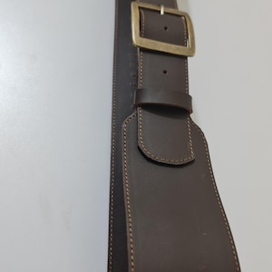 May include: A dark brown leather belt with a brass buckle. The belt has a wide design with decorative stitching along the edges. The buckle is rectangular and has a classic style. The belt is likely for a guitar or other instrument.