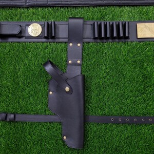 May include: Black leather gun holster and belt set. The belt features a gold-colored buckle, a decorative medallion, and several cartridge holders. The holster is designed to be worn on the hip and is secured with a strap.