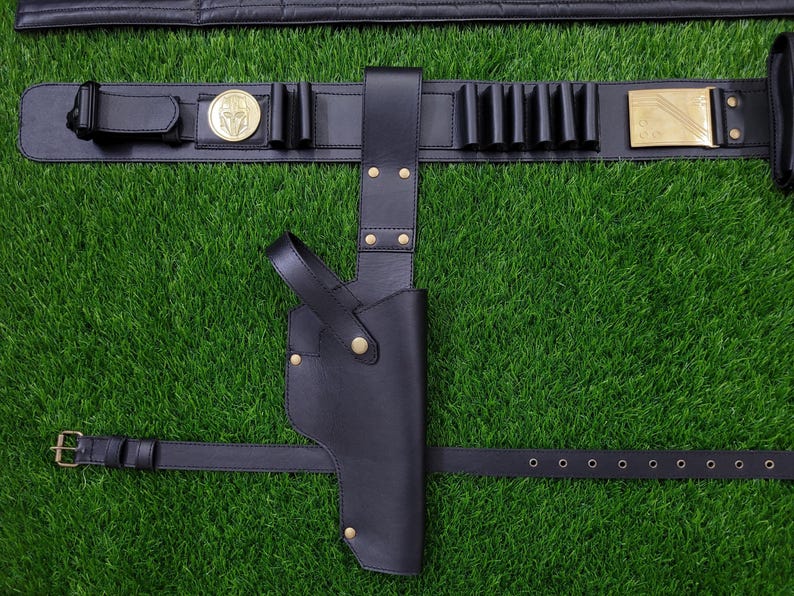 May include: Black leather gun holster and belt set. The belt features a gold-colored buckle, a decorative medallion, and several cartridge holders. The holster is designed to be worn on the hip and is secured with a strap.