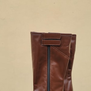 Puede incluir: Un par de botas de montar de cuero marr&oacute;n con una cremallera en la parte posterior y una correa en la parte superior.